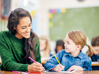Lehrerin und Kind sitzen nebeneinander an Schulbank und sehen sich an